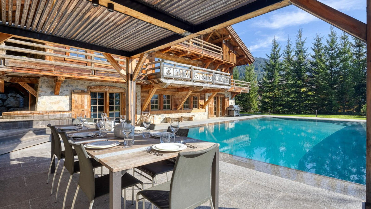 Outdoor dining area under a wooden pavilion overlooking a bright blue swimming pool, with a large log cabin and evergreen trees in the background.