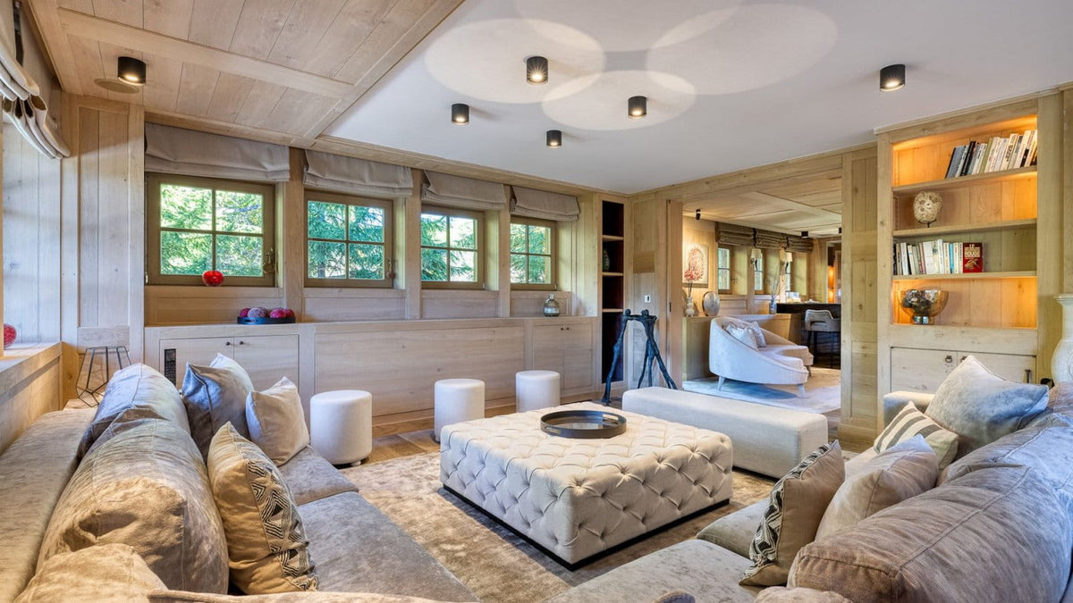 A luxury modern chalet living room featuring a tufted cream ottoman, plush beige sofas, white cylindrical stools, and light wood paneled walls and ceiling.