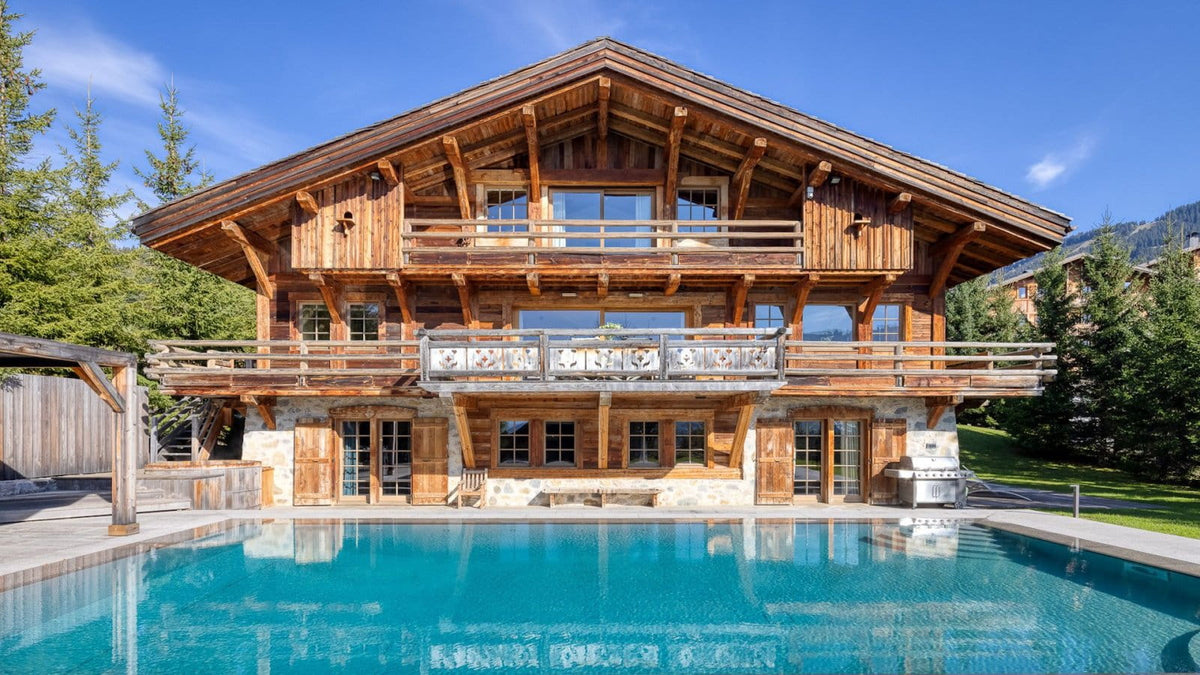 A large alpine-style wooden chalet with multiple balconies, a swimming pool in the foreground, trees, and blue sky.