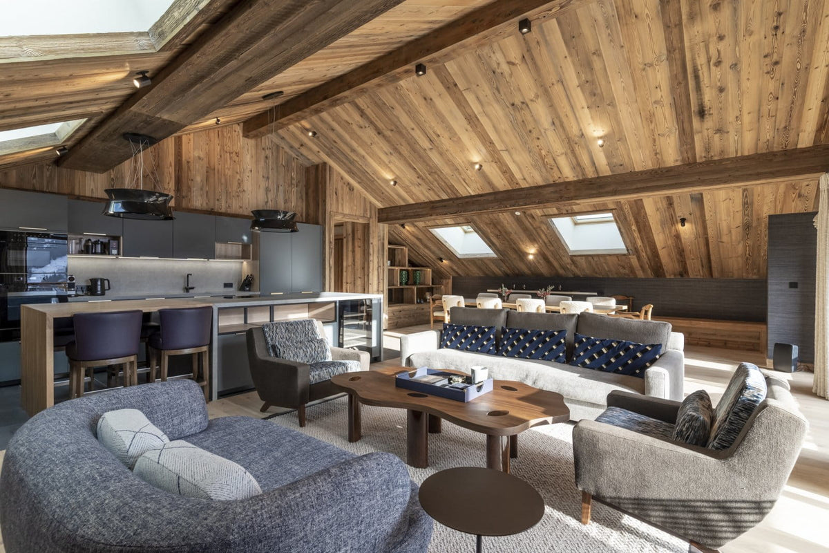 A spacious, modern ski chalet living room with a high wood-beamed vaulted ceiling, featuring a gray textured sofa in the foreground, a white sofa with blue patterned cushions, a wooden coffee table, and a kitchen area in the background.