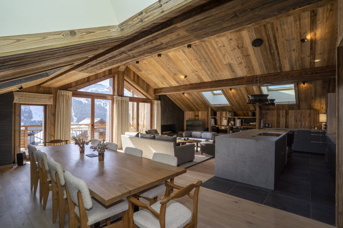 A large, modern chalet-style dining and living room with exposed wood ceiling beams, a wooden dining table with upholstered chairs, a gray kitchen island, and wide windows looking out onto a mountain view.