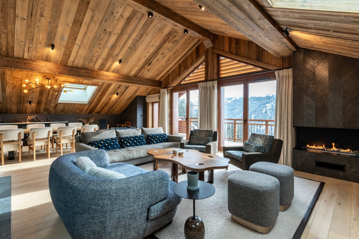 A luxurious ski lodge living room with a high wood-beamed ceiling, featuring a gray sectional sofa, two blue-gray armchairs, matching ottomans, a wooden coffee table, and a built-in fireplace.