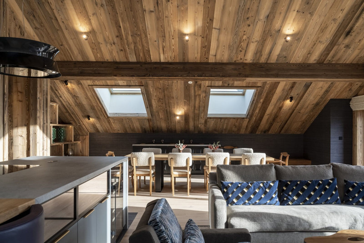 A modern ski chalet living room with a gray sofa, blue patterned pillows, a wooden dining area, and a slanted wooden ceiling with skylights.