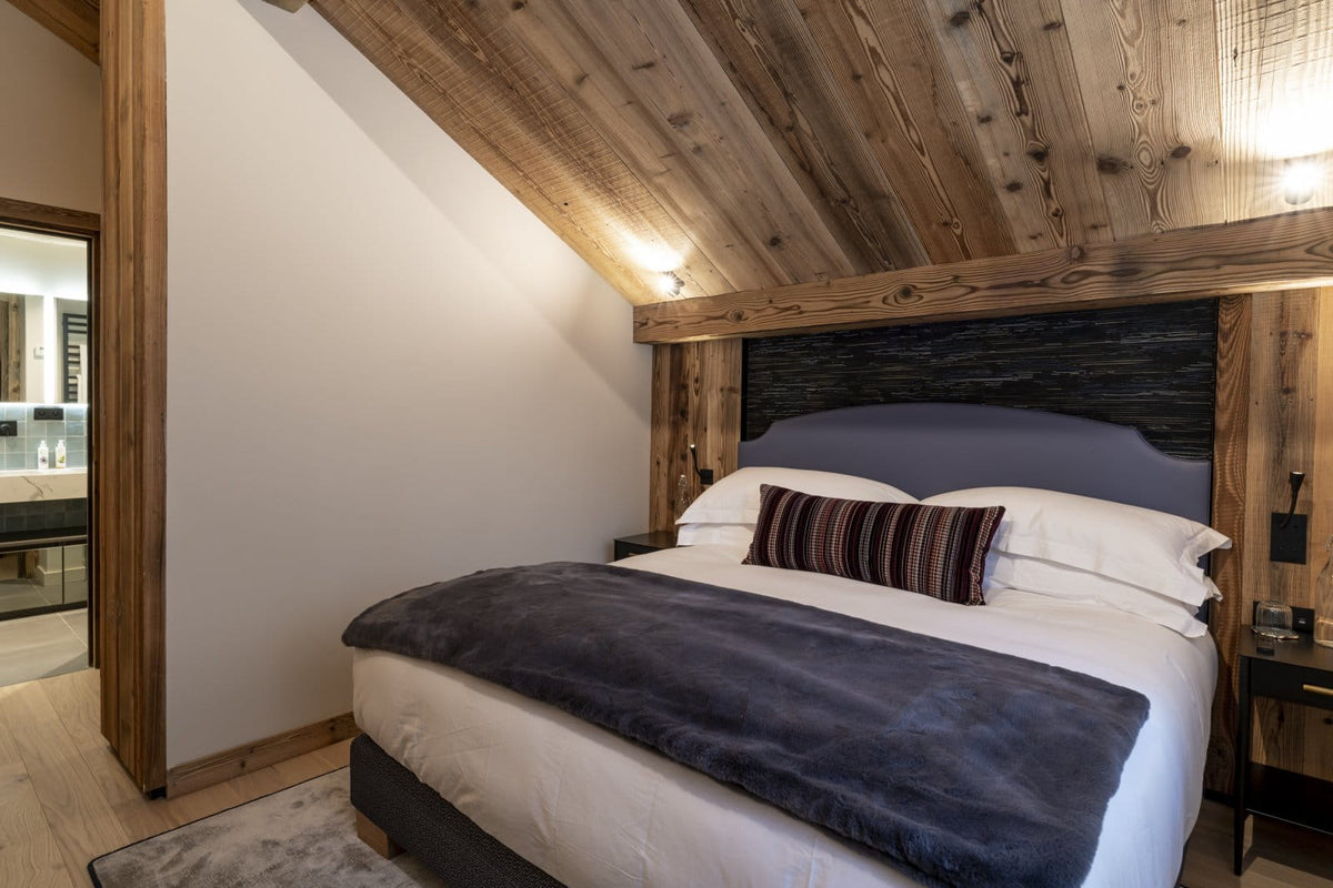 A cozy bedroom with a bed dressed in white linens, a dark gray throw blanket, and a striped accent pillow, set against a rustic wooden headboard and matching wood-paneled ceiling.