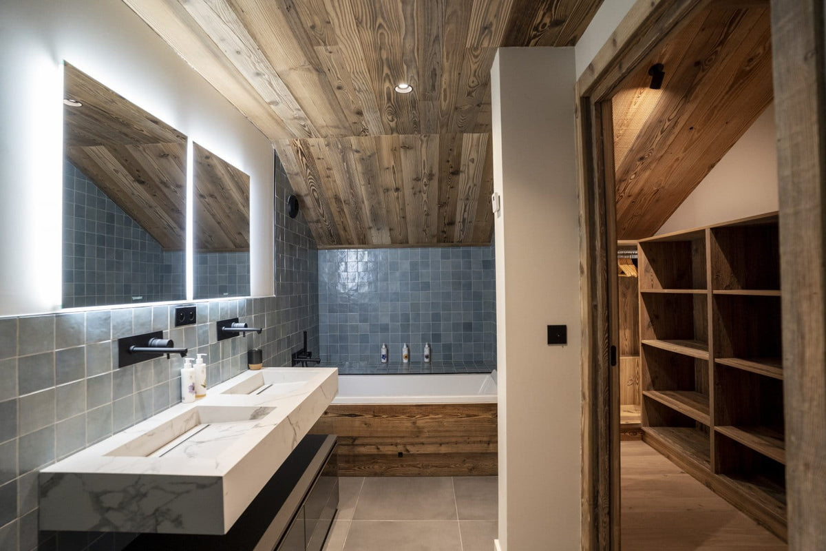 A modern bathroom interior with a double sink vanity, large backlit mirrors, blue-gray tiled walls, a wooden bathtub, and an open doorway revealing built-in wooden shelving.
