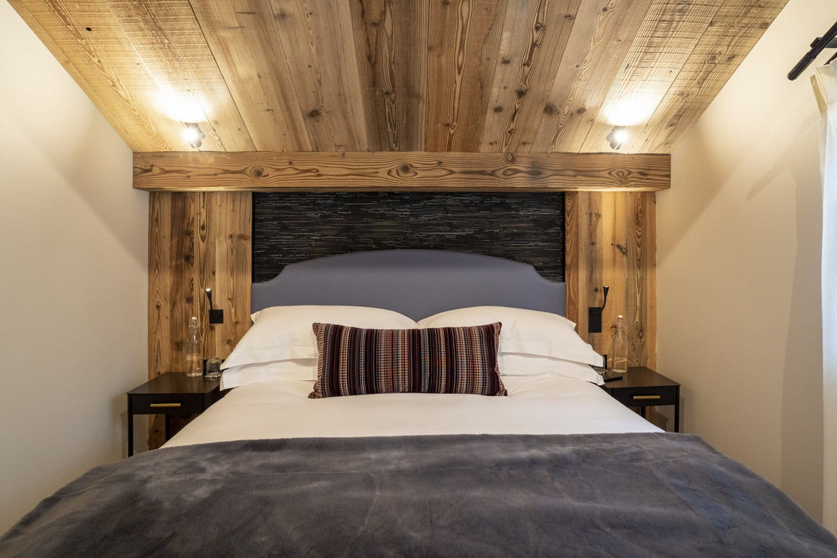 A rustic bedroom setup featuring a bed with white linens and a striped accent pillow, set against a dark wood headboard and framed by a large wooden beam under a wood-paneled ceiling with wall-mounted lights.