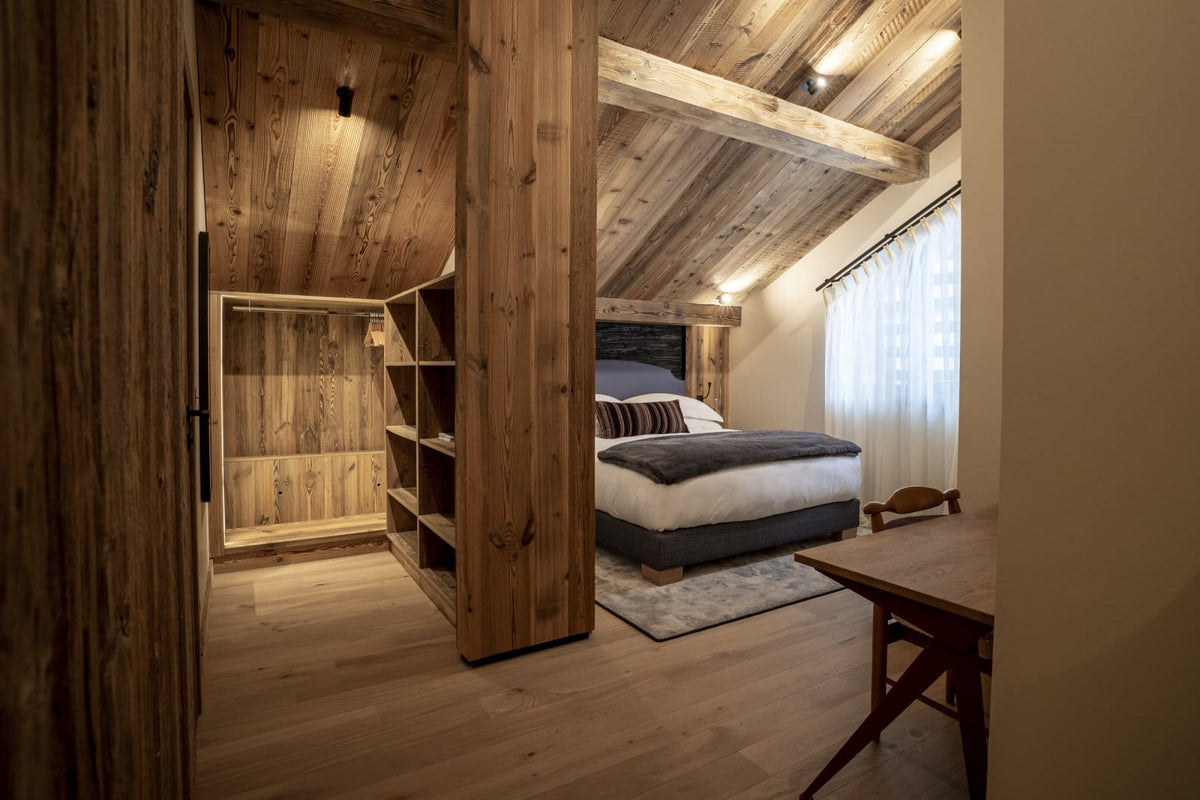 A rustic bedroom interior with a wooden slatted partition, built-in shelving, a bed with white and gray bedding, a wooden table, and warm wood ceiling and floor panels.