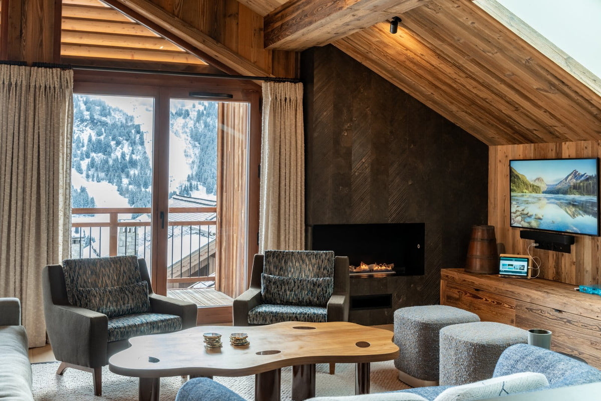 A cozy ski chalet living room with a wood-burning fireplace, two gray armchairs, a curved wooden coffee table, and a wall-mounted TV showing a mountain landscape. Large windows offer a view of snowy mountains outside.