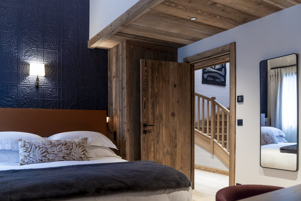 A rustic bedroom interior with a large bed, dark textured wall, wooden ceiling beams, an open wooden door revealing a staircase, and a wall mirror.