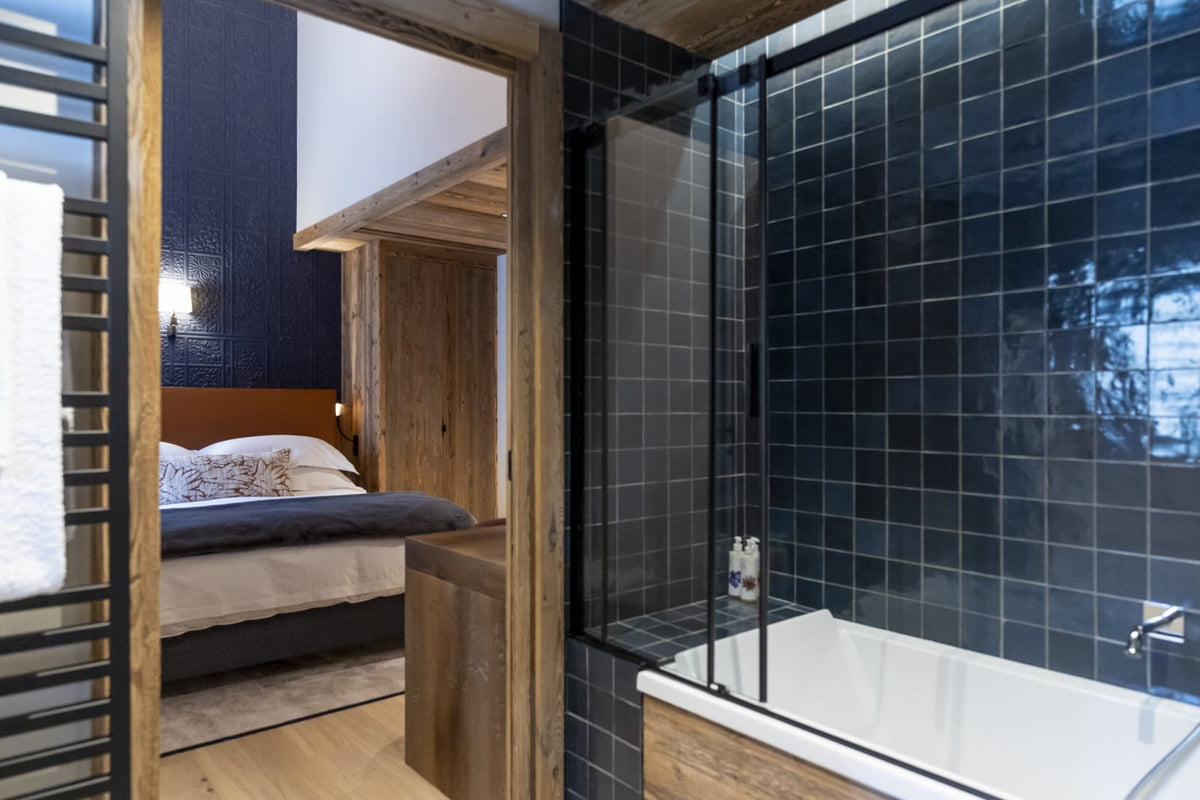 Modern bathroom interior with a white bathtub, dark blue tiled wall, glass shower enclosure, and an open doorway revealing a wooden bedroom with a bed.