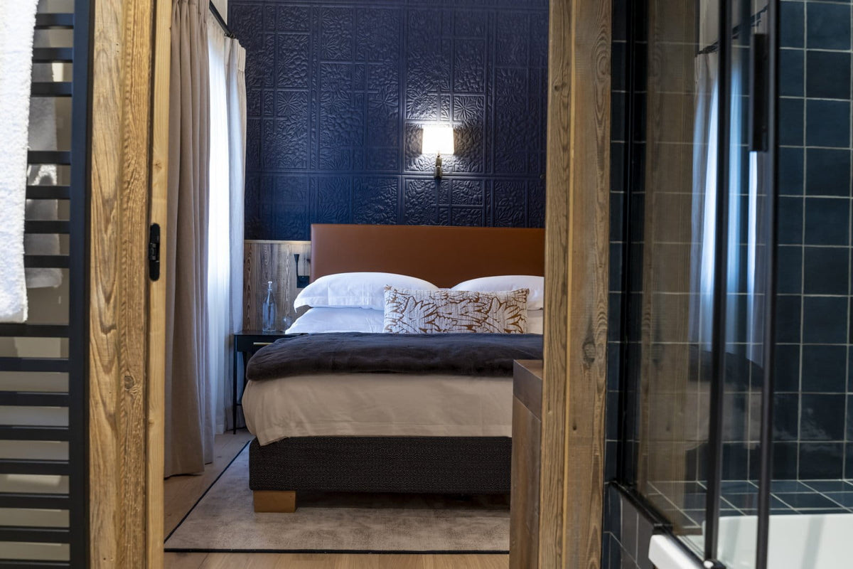 A modern bedroom interior viewed through an open wooden doorway, featuring a bed with white and dark gray bedding, a dark blue textured wall, a wall-mounted light, and a glass shower enclosure on the right.