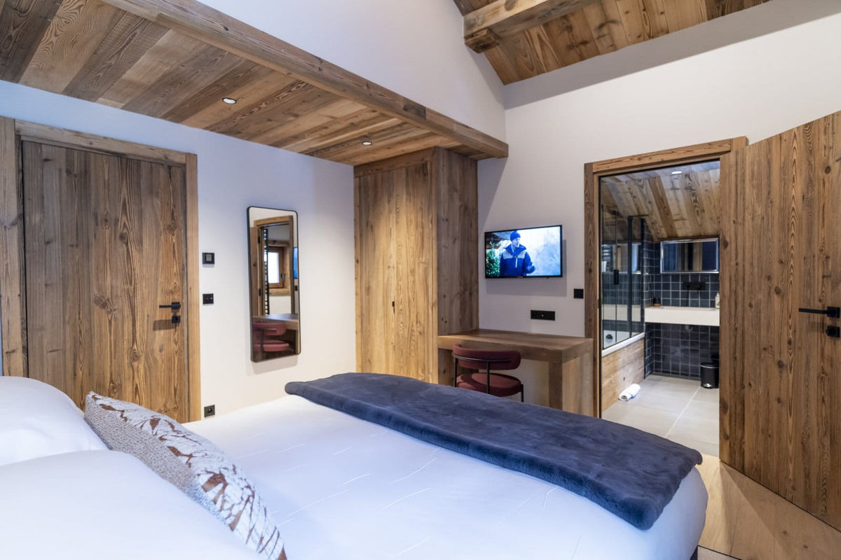 A modern rustic bedroom featuring a large bed with white bedding and a dark blue throw blanket, set in a room with prominent wooden ceiling beams, a wooden desk, and a built-in TV. An open wooden door on the right reveals a tiled bathroom.