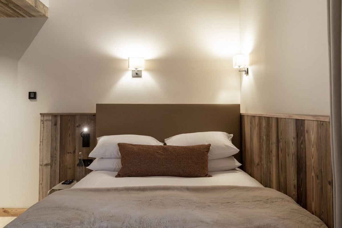 A neatly made bed in a rustic bedroom with a brown upholstered headboard, wood-paneled side walls, two wall-mounted sconces, white bedding, and a brown accent pillow.