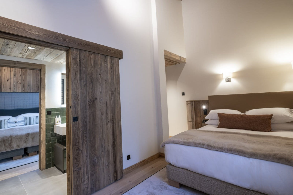 A modern bedroom interior featuring a large bed with white bedding and a brown pillow, next to a rustic dark wood sliding door that opens into a bathroom. The room has warm neutral walls, wooden ceiling details, and soft ambient lighting.