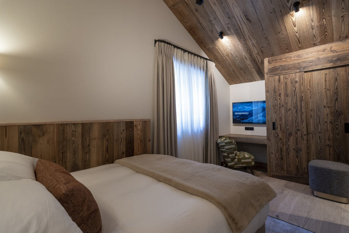 A cozy bedroom interior with a neatly made bed, warm wood-paneled walls and ceiling, sheer curtains, a small desk area with a TV, and a gray upholstered ottoman in a rustic mountain-style room.