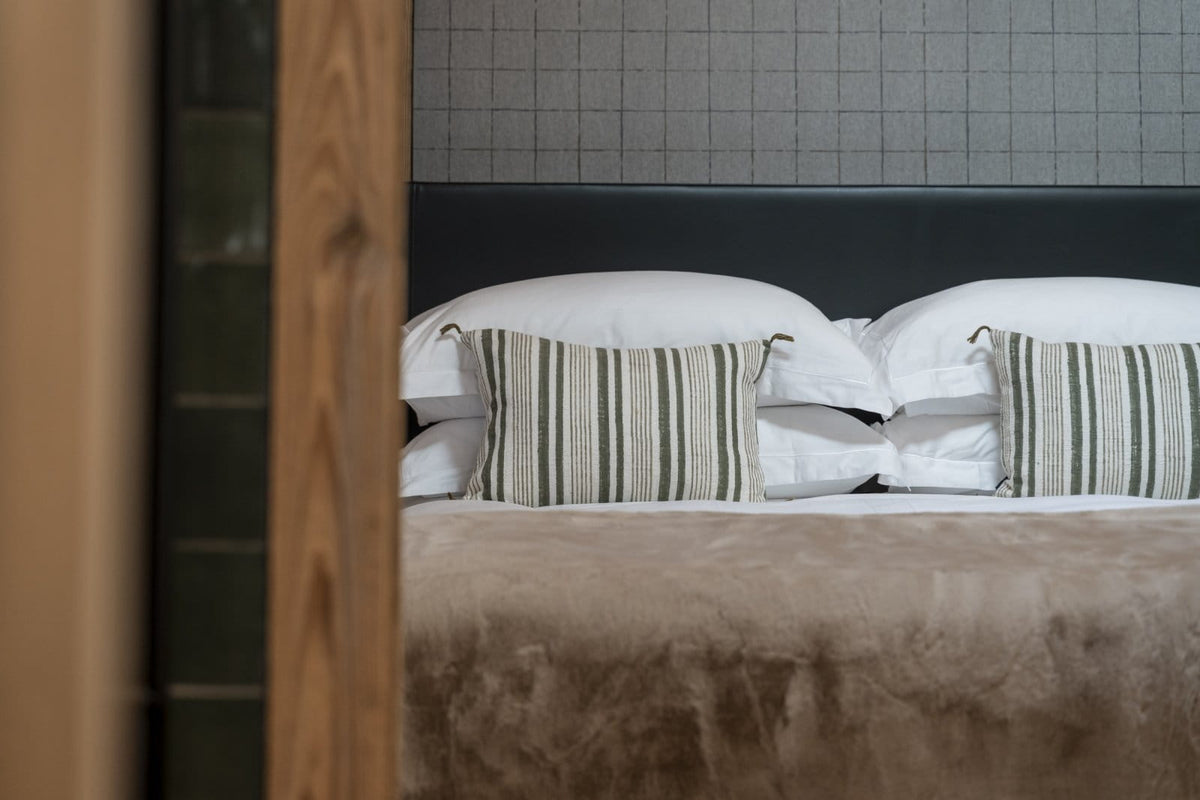 A close-up view of a neatly made bed with white pillows, two striped accent pillows in beige and green, a brown headboard, and a textured wall behind it. A wooden frame is visible in the foreground on the left.