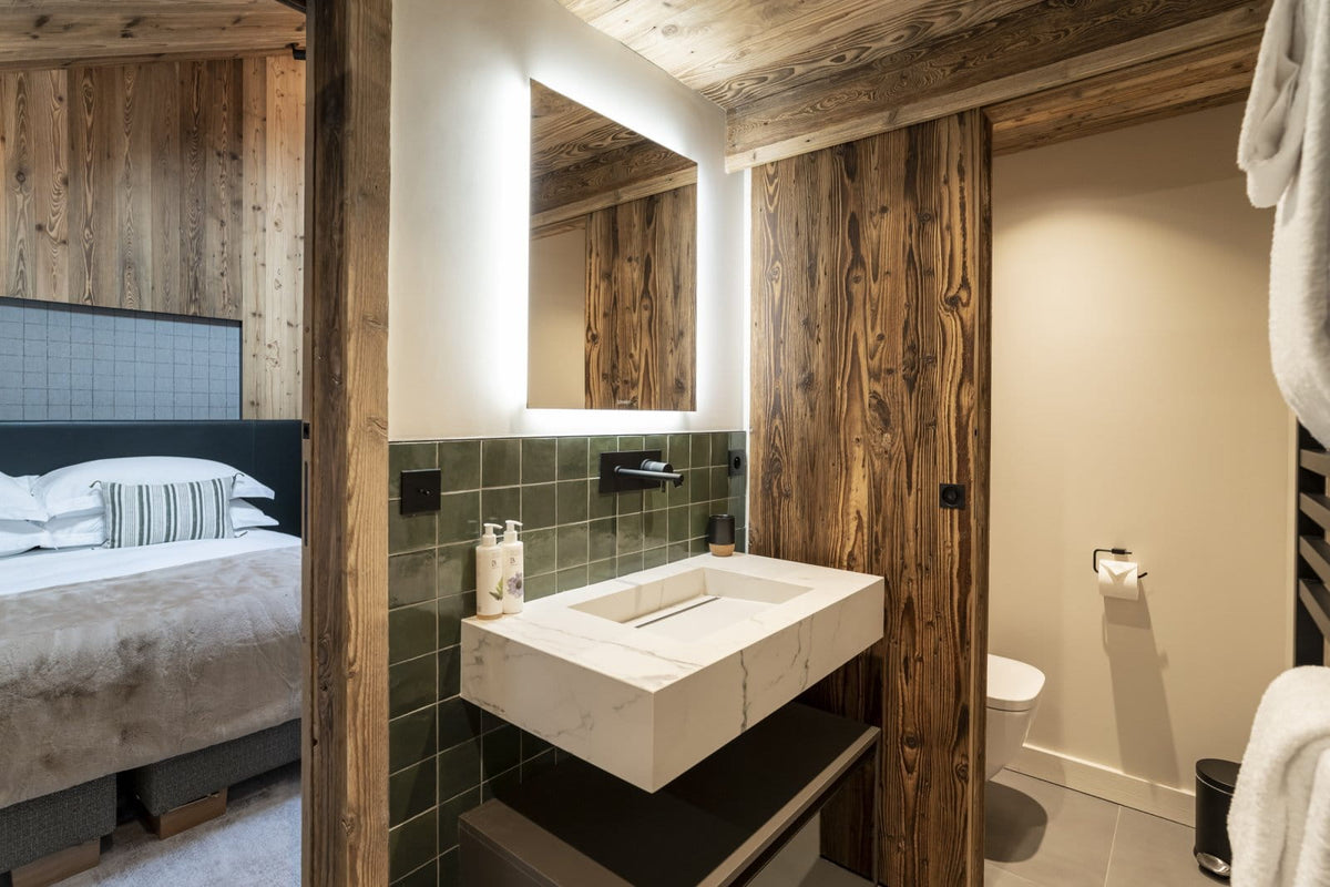 A modern rustic bathroom featuring a floating white stone sink, a large backlit rectangular mirror, dark green wall tiles, and prominent dark wood paneling and beams. A white towel is hanging on the right, and a glimpse of a bed is visible on the left.