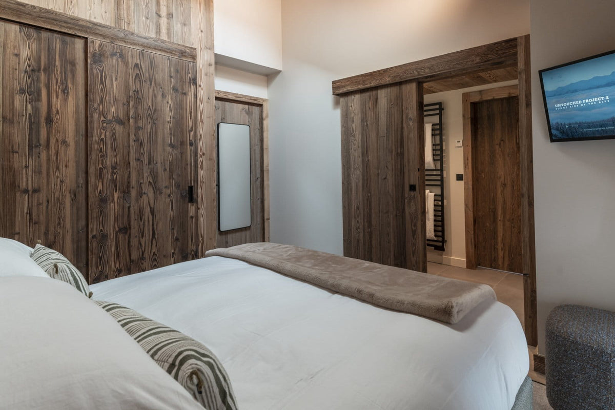 A modern bedroom interior featuring a large bed with white bedding and a brown throw blanket, set against rustic dark wood paneling and sliding doors. A wall-mounted TV is visible on the right wall.