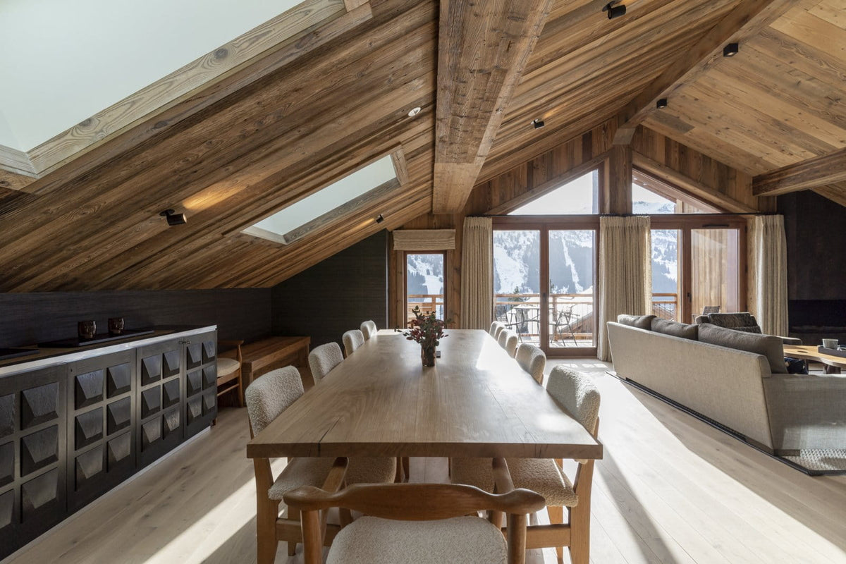 A luxury ski chalet interior featuring a long wooden dining table and chairs, a modern kitchenette, a large living room with a sofa, and a high-sloped wooden ceiling with skylights looking out to mountains.