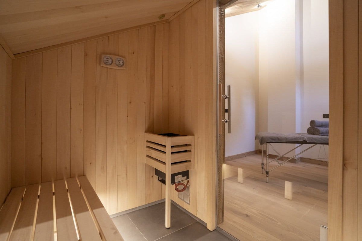 A wooden sauna interior with a built-in wooden sauna bench, a wall-mounted wooden steam basket, and an open door revealing a spa treatment room with a massage table.