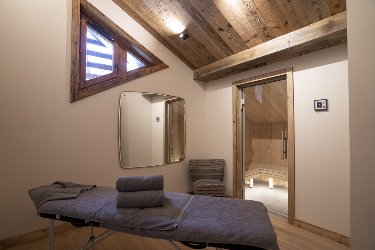 A spa treatment room with a massage table covered in gray towels, a wall mirror, a small chair, and a wooden sauna visible through an open door. The room has warm wood ceiling beams, light walls, and a window with wooden framing.