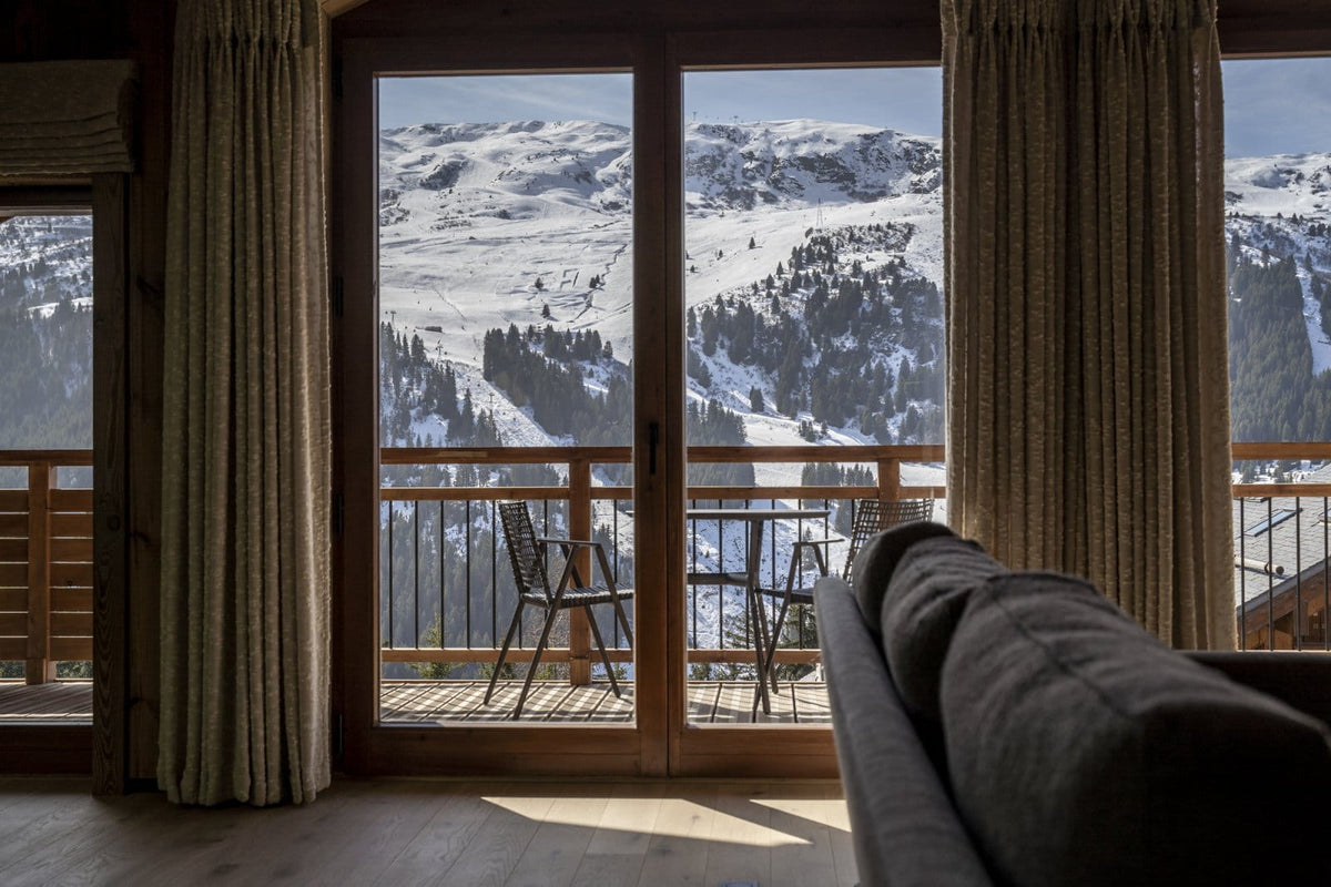 A cozy interior room with large floor-to-ceiling windows overlooking a snowy mountain landscape. The room has wood-framed doors, beige curtains, a wooden railing, and part of a dark gray sofa in the foreground.