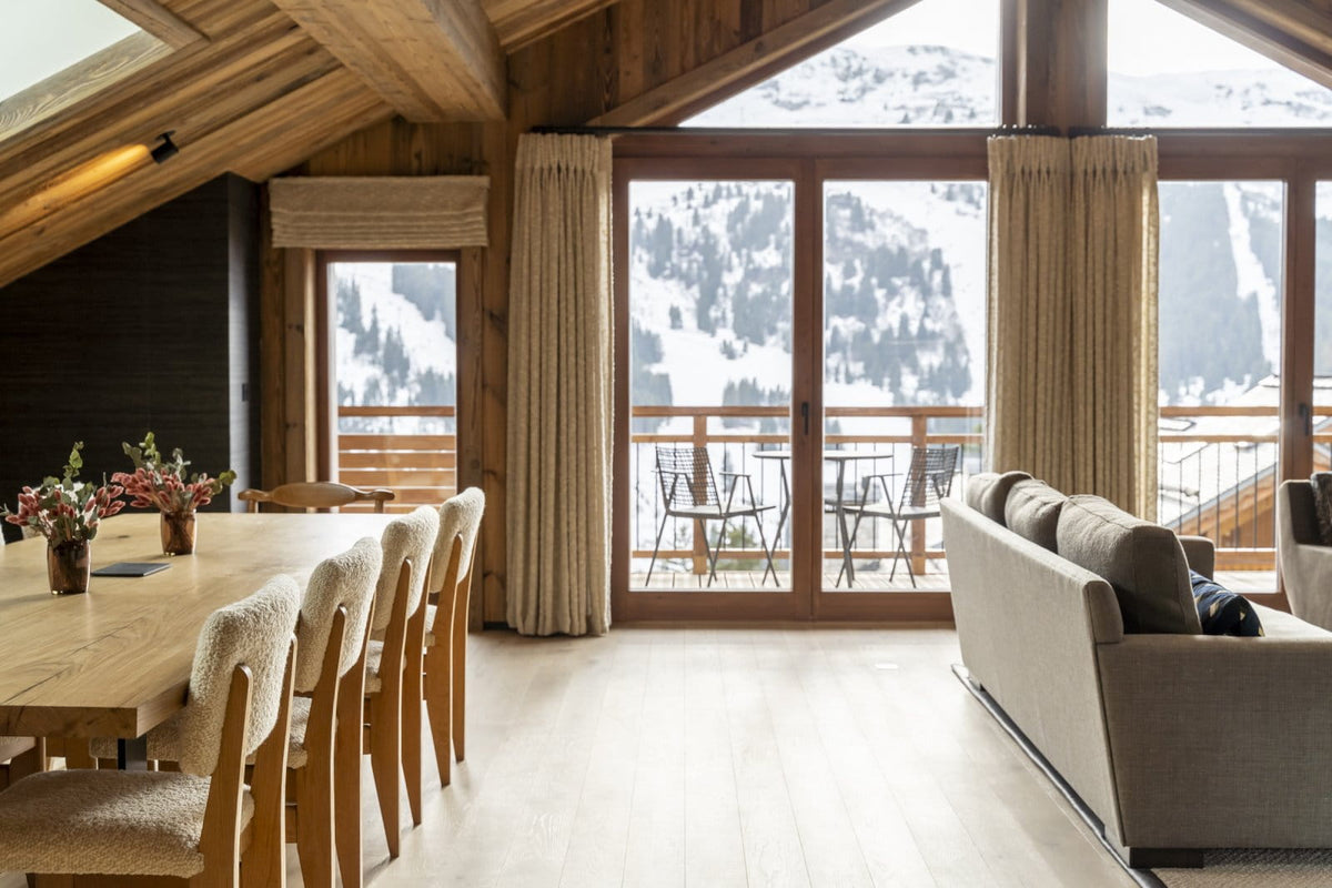 A bright, modern chalet-style living and dining room with large floor-to-ceiling windows overlooking a snowy mountain landscape. The interior features warm wood walls, a light wood dining table with upholstered chairs, beige curtains, and a light gray sofa.