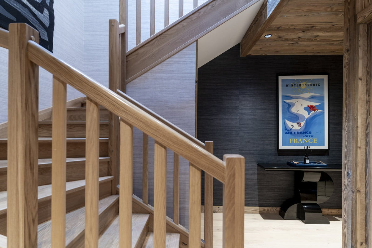 A modern wooden staircase with light oak railings and steps, leading up an interior level. The background features a dark gray textured wall with a framed vintage-style travel poster and a black console table.
