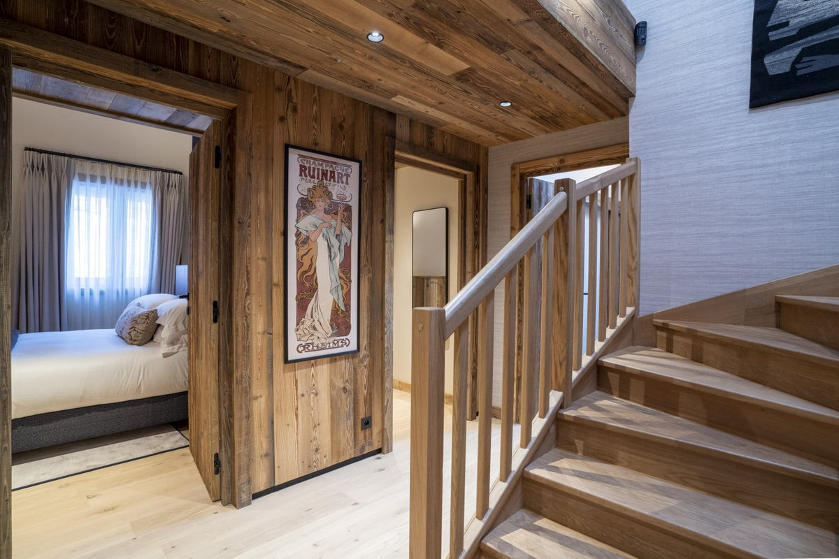 A rustic wooden staircase with a light wood handrail in a modern chalet-style apartment, featuring a bedroom visible through an open doorway on the left.