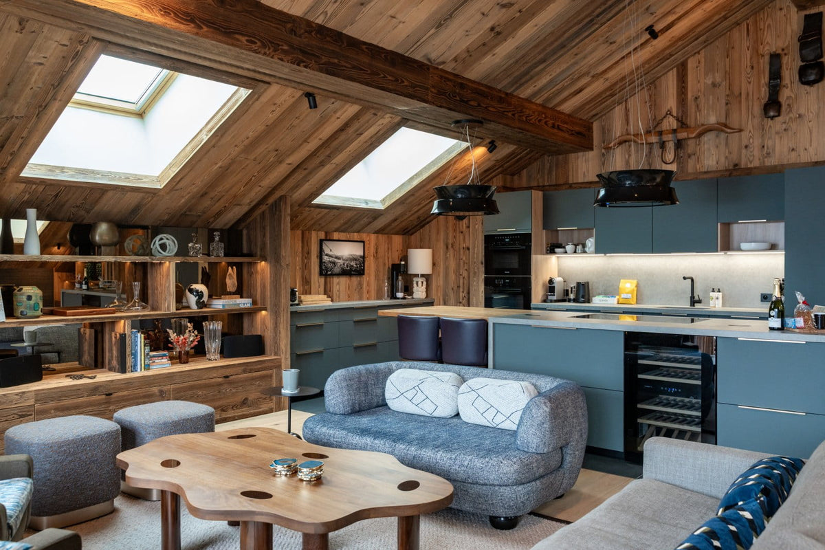 A luxury modern chalet interior featuring a large gray fabric sofa, a rustic wooden coffee table, a kitchen with slate blue cabinets, and a sloped wooden ceiling with skylights.