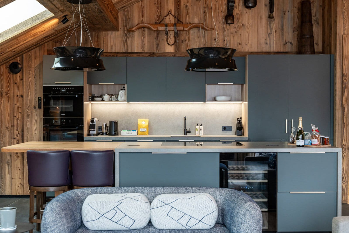 A modern kitchen island with dark gray cabinets, a light wood countertop, two dark purple bar stools, and a gray sofa in the foreground, set within a rustic wood-paneled room.