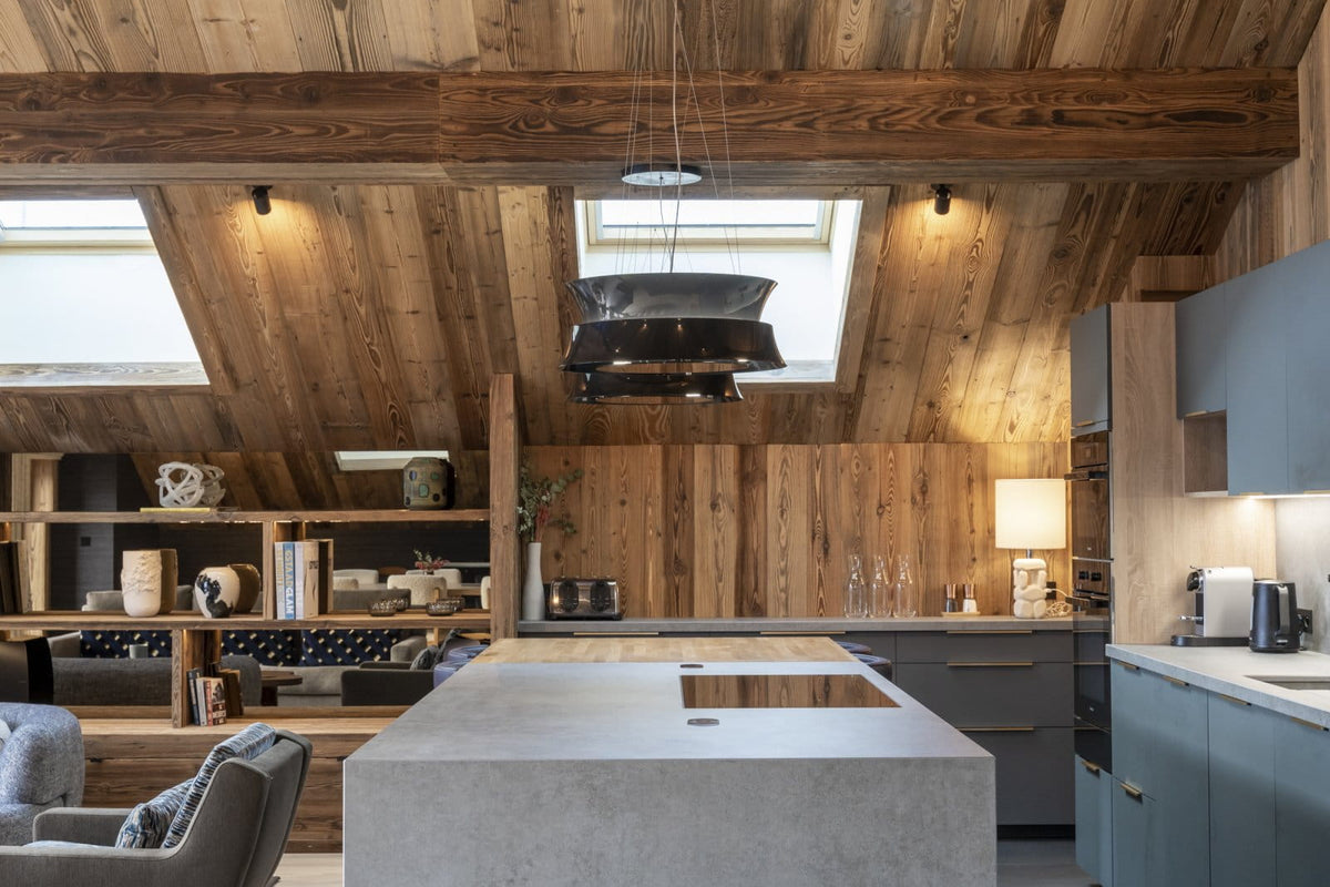 A modern kitchen interior with a large concrete island in the foreground, wood-paneled walls and ceiling, built-in cabinetry, shelving, and a pendant light hanging from the ceiling.