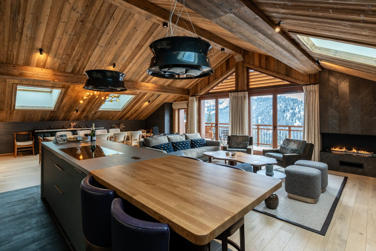A luxury ski chalet interior featuring a large wooden kitchen island with dark bar stools, a modern living room with a fireplace, and a high wood-paneled ceiling with skylights.