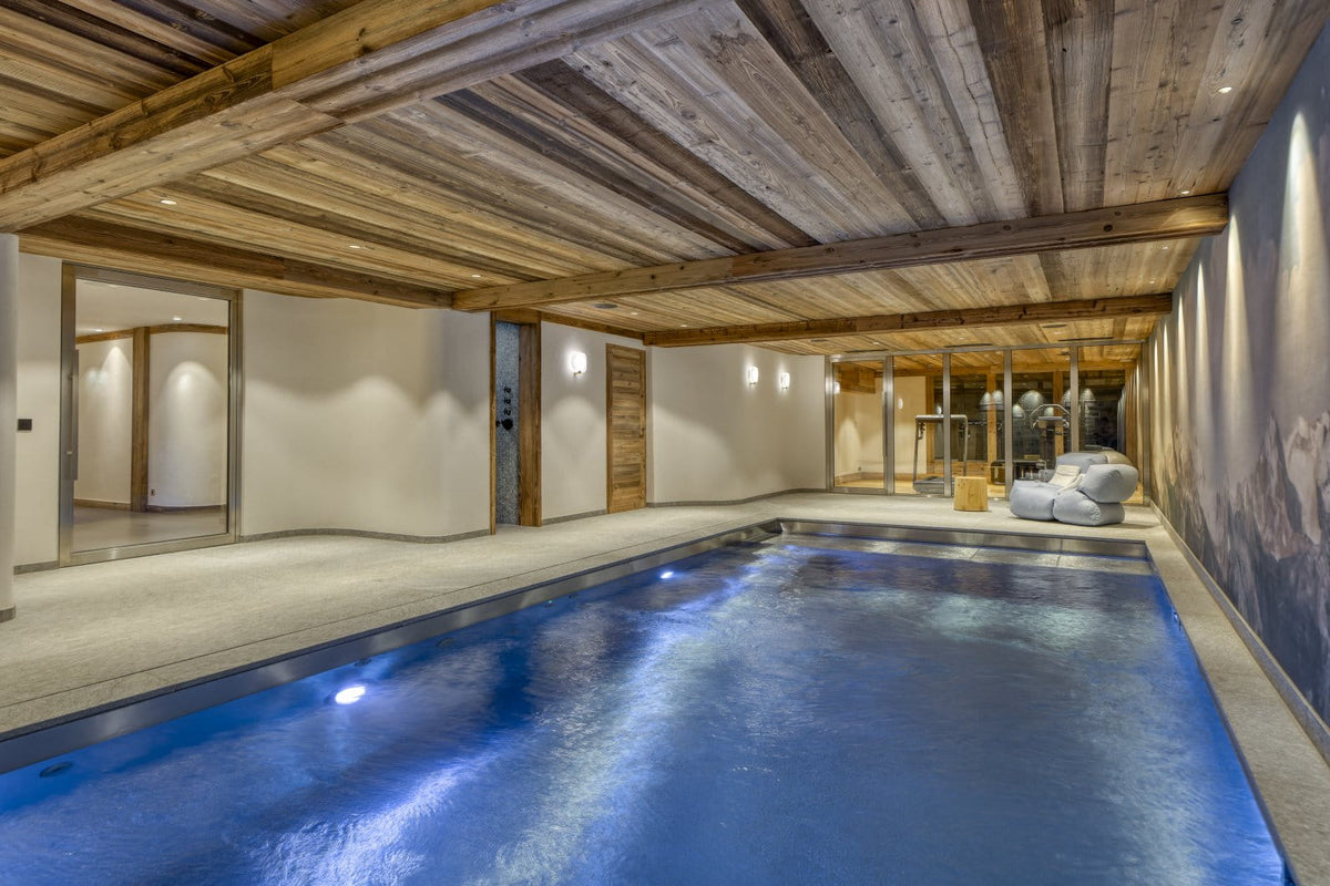 An indoor swimming pool with blue water, surrounded by light-colored walls and a rustic wooden ceiling with exposed beams.