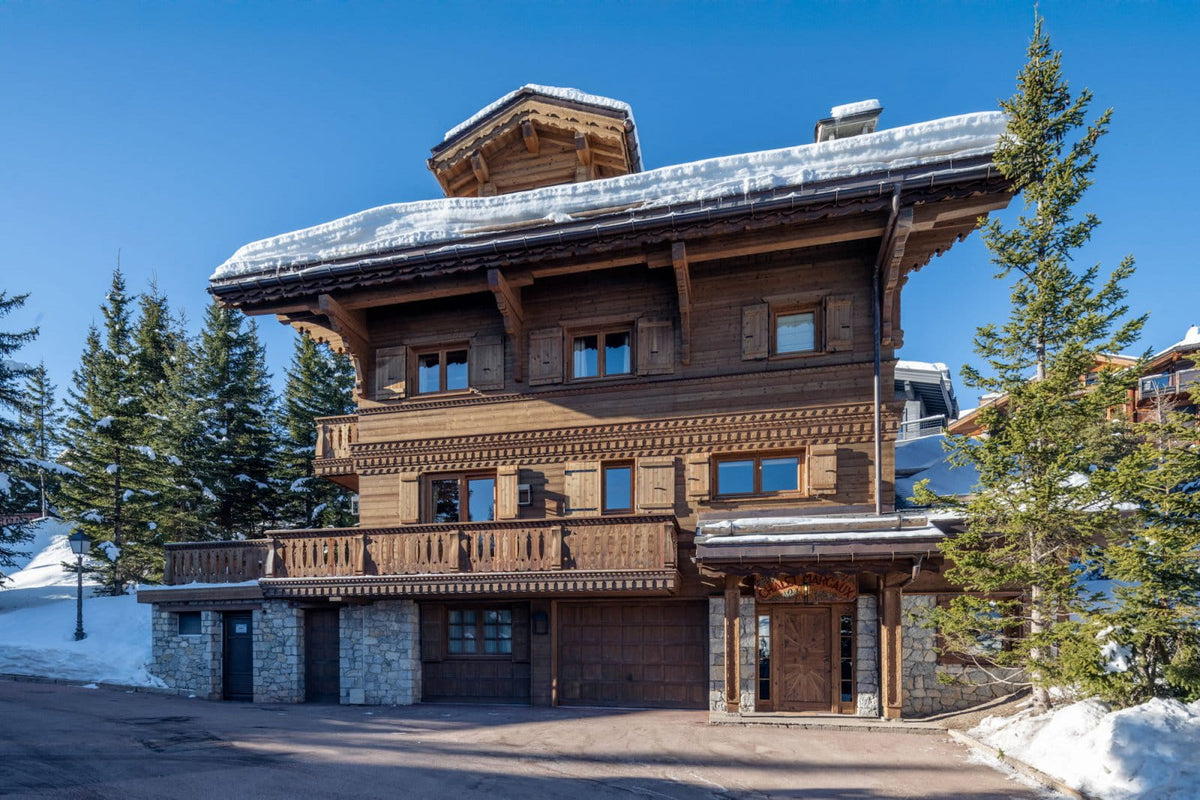 A large traditional alpine chalet with dark wood balconies and snow-dusted roofs, surrounded by evergreen trees and snow under a clear blue sky.