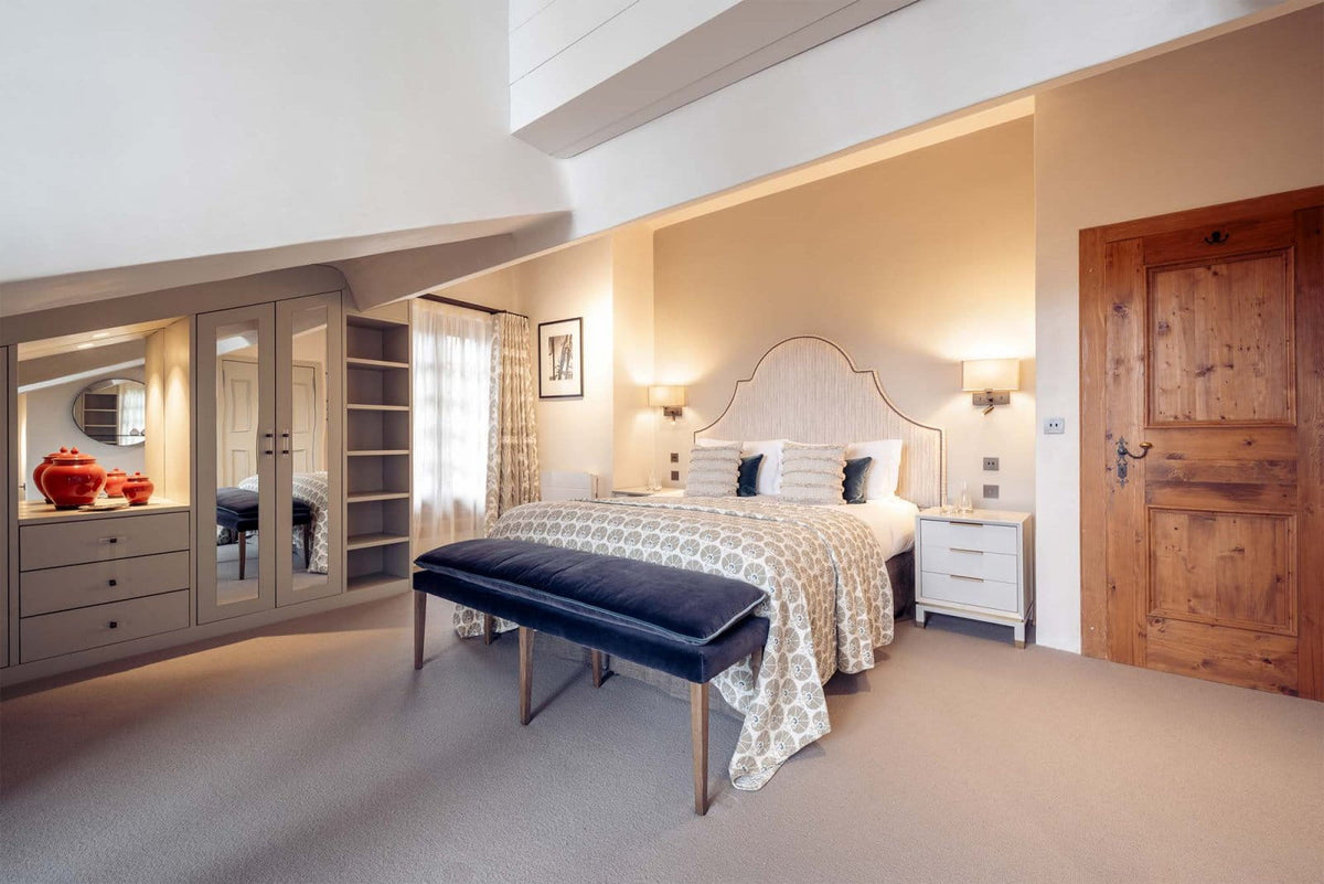 A styled bedroom interior featuring a bed with patterned beige and white bedding, a dark blue upholstered bench at the foot, built-in wardrobes with mirrors, warm wall lighting, and a rustic wooden door on the right.
