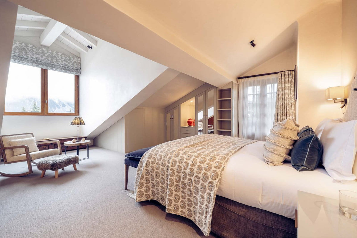 A modern slanted-roof bedroom featuring a large bed with white linens and a beige patterned throw blanket, a matching armchair, a window with a patterned roller blind, and built-in shelving.