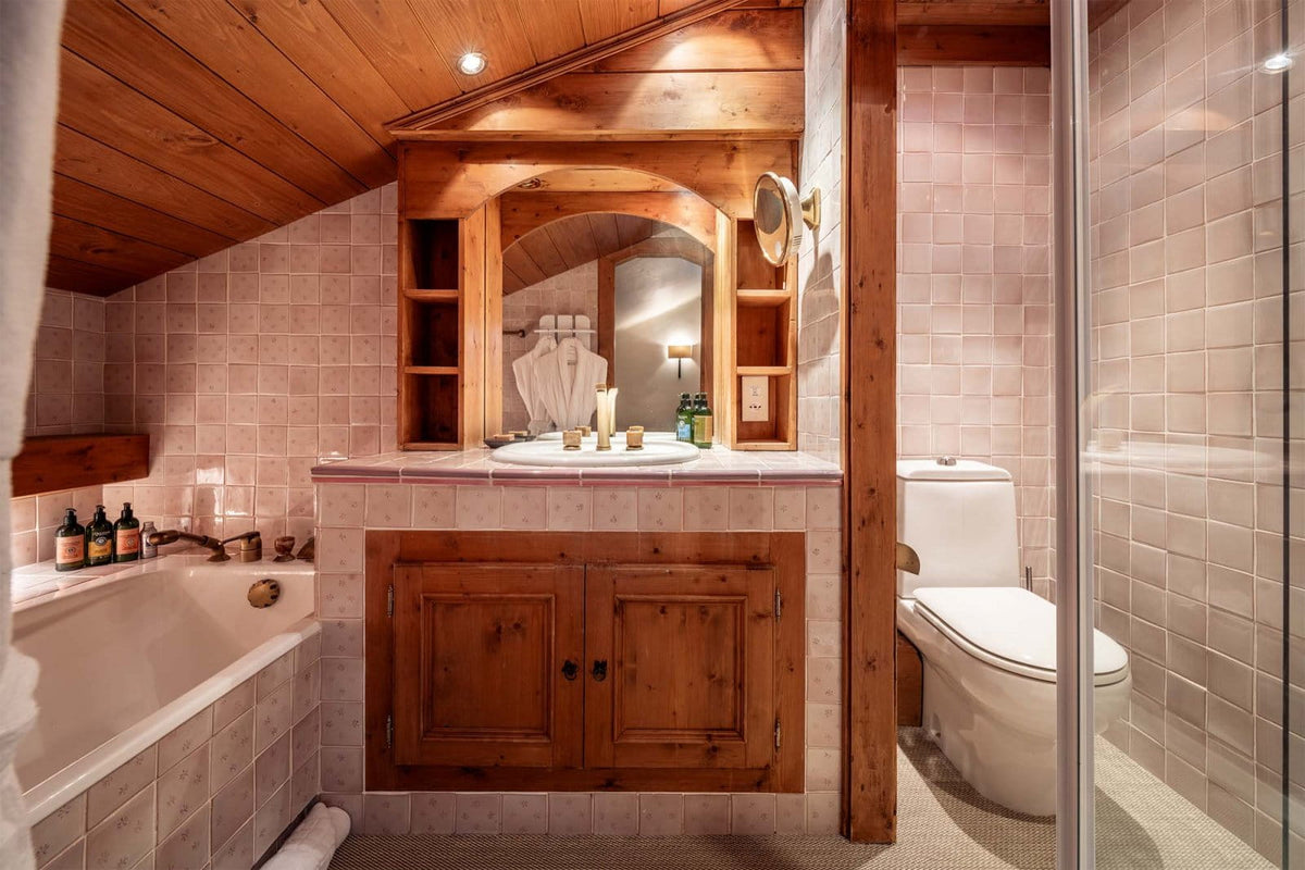 A rustic bathroom interior with a wooden vanity cabinet, arched mirror, tiled walls, a bathtub on the left, a toilet on the right, and a glass shower enclosure. The room has warm wood ceiling panels and light beige tile surfaces.