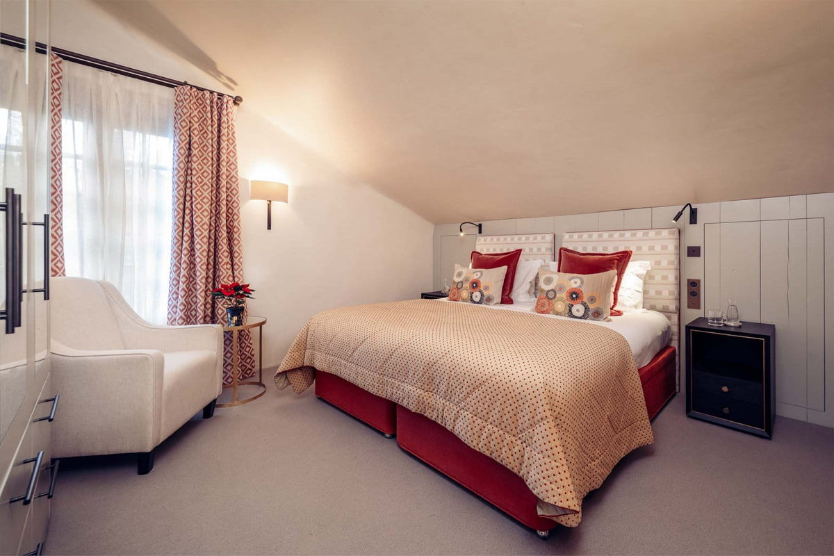 A cozy bedroom scene featuring a bed with a beige patterned quilt, red bed frames, and red pillows, alongside a white armchair, patterned curtains, and a bedside table.