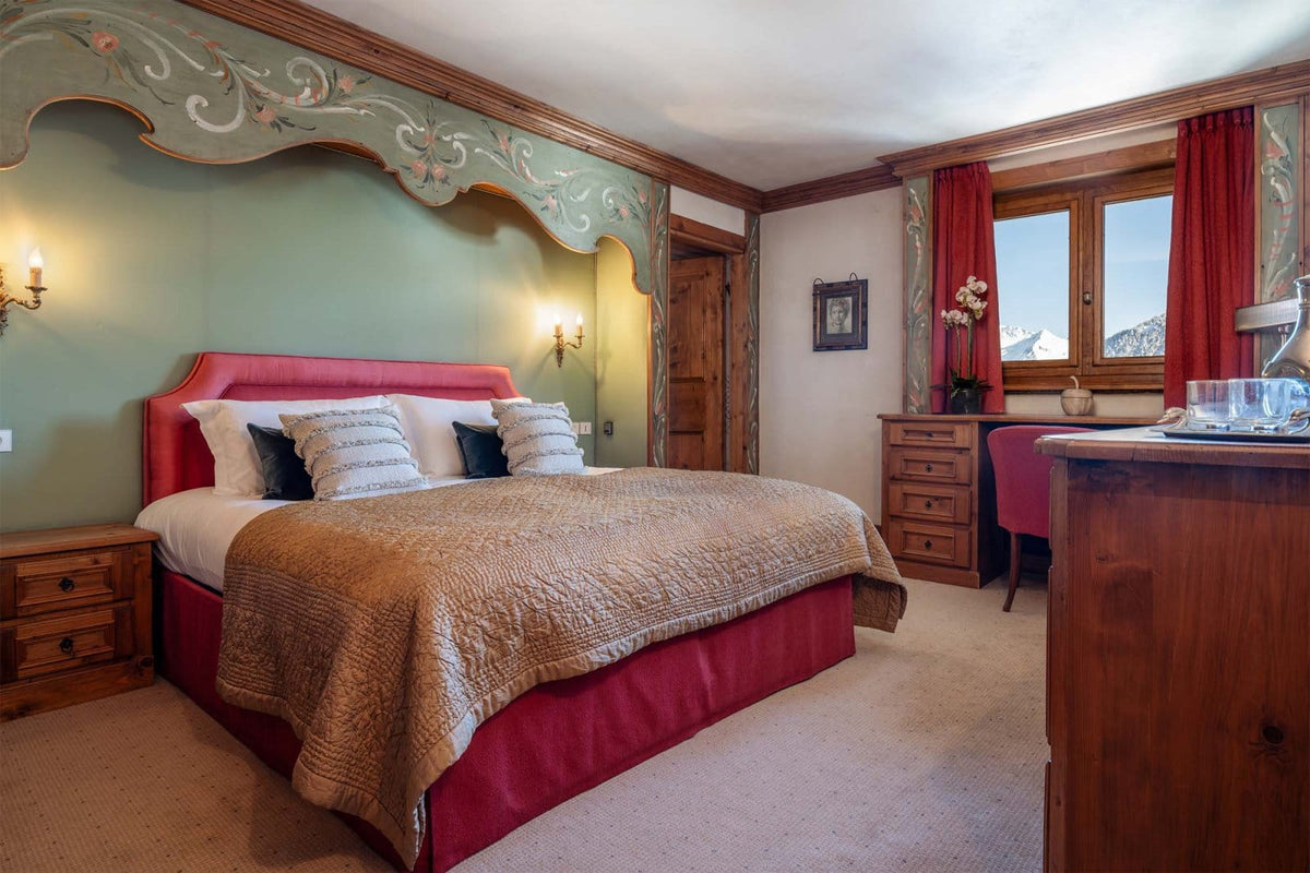 A cozy alpine-style bedroom featuring a bed with a red upholstered headboard and skirt, a beige quilted blanket, decorative pillows, a wooden dresser, and a window with red curtains looking out to a mountain view.