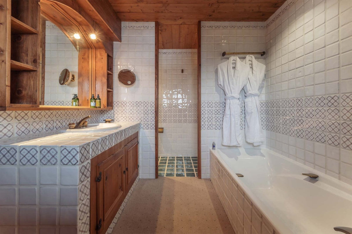 A rustic mountain-style bathroom featuring a white tiled bathtub with a white bathrobe hanging on the wall, a matching vanity with wooden cabinets and a large mirror, and a walk-in shower area in the background.