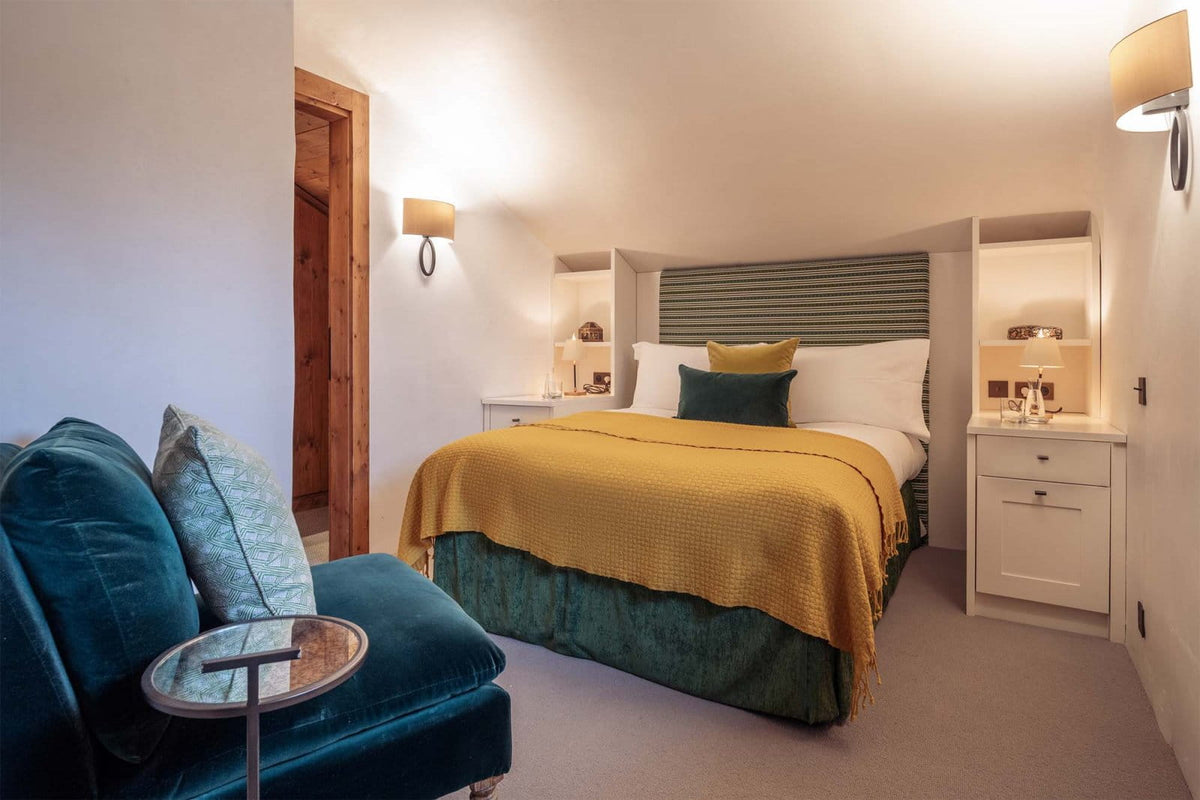 A cozy bedroom interior featuring a bed dressed in white linens with a mustard yellow textured throw and dark green pillows, alongside a teal velvet armchair in the foreground and built-in bedside cabinets with warm lighting.