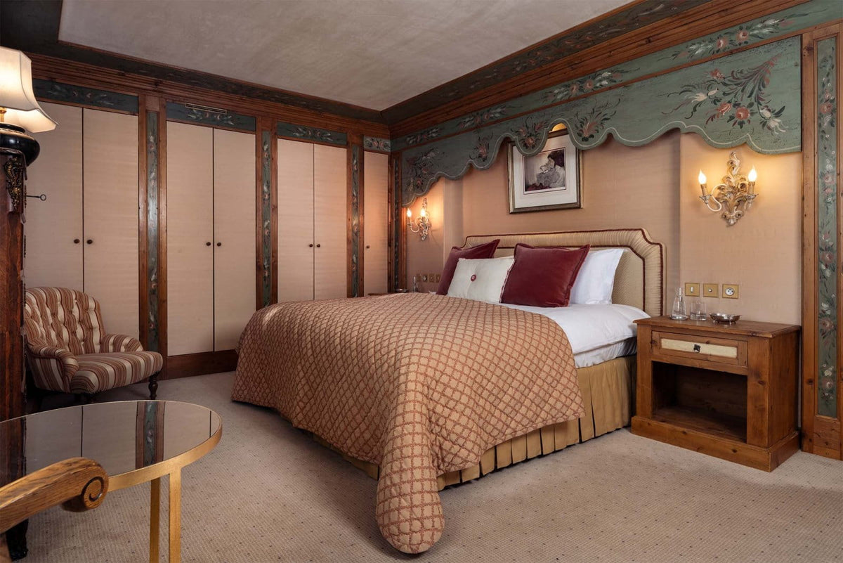 A classic bedroom interior featuring a bed with a beige and brown checkered quilt, white pillows, and deep burgundy accent pillows. The room has warm wood paneling, built-in wardrobes, a striped armchair, and wall sconces.