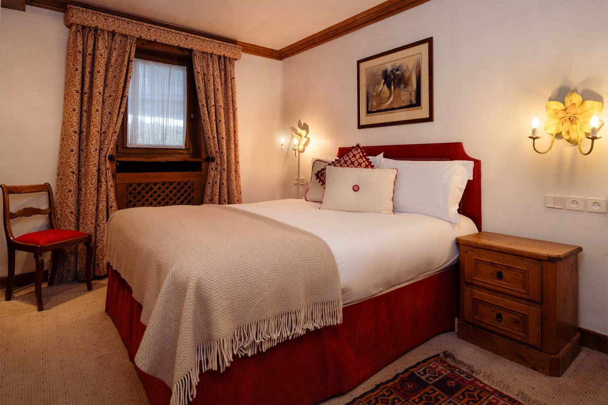 A cozy bedroom interior featuring a double bed dressed in white linens with a red bed skirt and red headboard, a beige fringed throw blanket, patterned curtains, a wooden nightstand, and wall-mounted flower-shaped lamps.