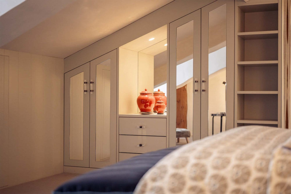 A built-in wardrobe or closet system in a bedroom, featuring cream-colored doors with glass panels, a central drawer unit, open shelving, and warm ambient lighting.