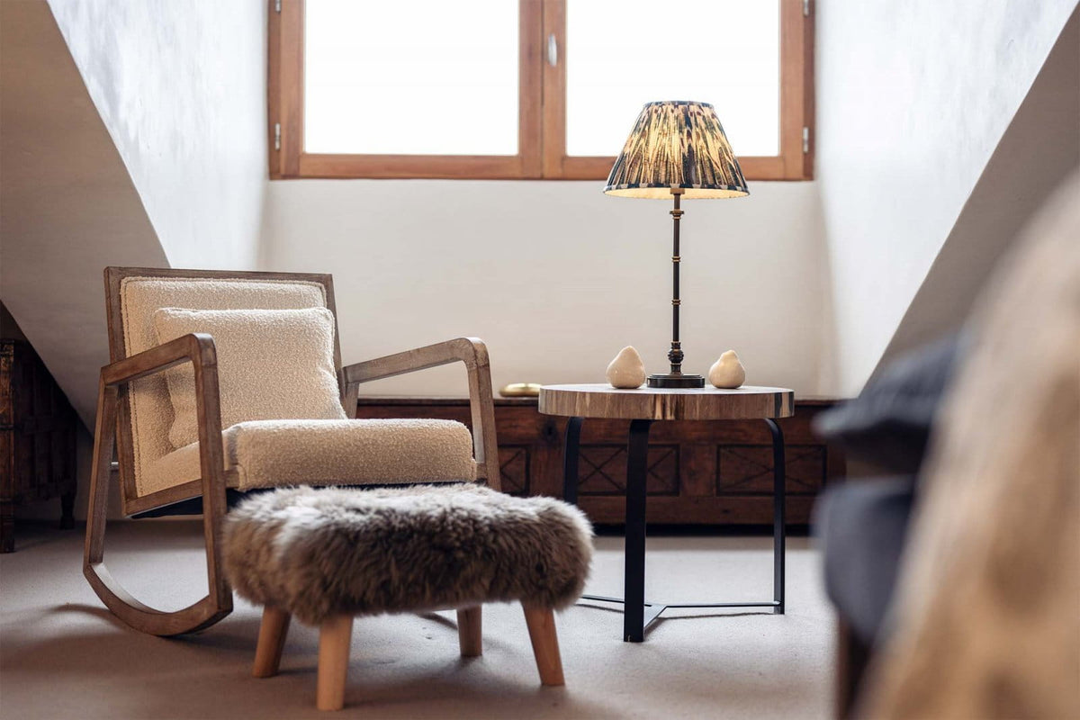 A cozy interior scene with a wooden-framed rocking chair upholstered in cream fabric, a matching furry ottoman, a small round side table with a lit lamp, and a window in the background.