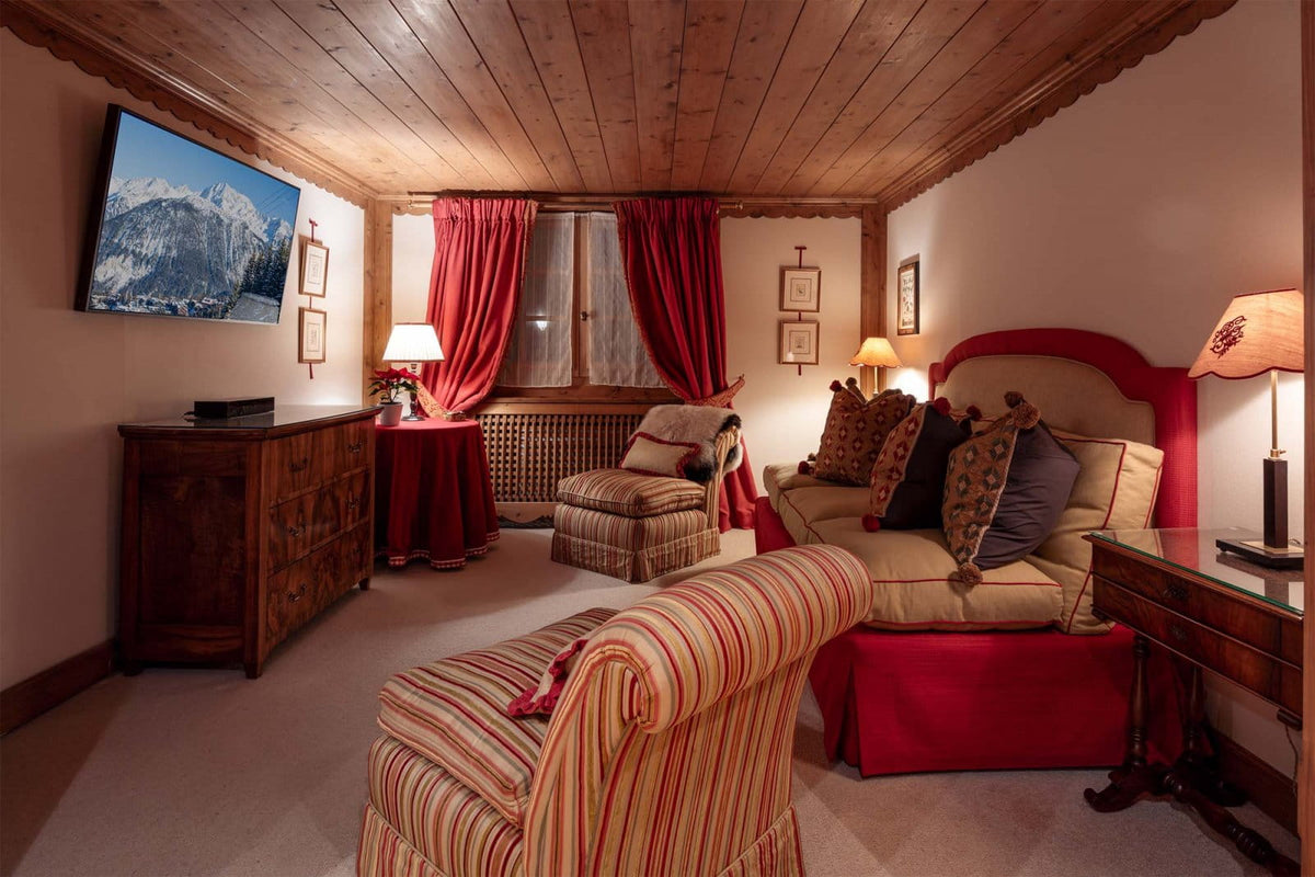A cozy mountain-style bedroom with a large bed dressed in red and beige bedding, matching striped armchair in the foreground, red curtains, wood-paneled ceiling, and a wall-mounted landscape TV.