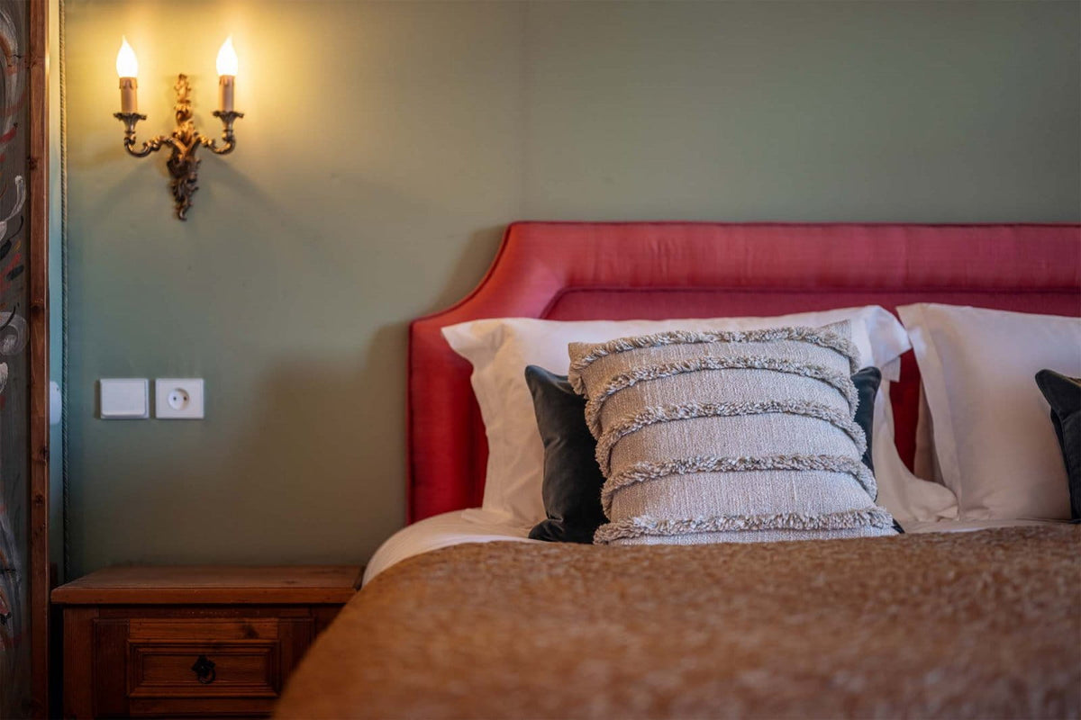 A close-up of a bed in a bedroom setting, featuring a prominent red upholstered headboard, white pillows, a textured beige throw blanket, and a central decorative pillow with horizontal metallic stripes. A warm wall-mounted sconce is visible on the olive-green wall to the left.