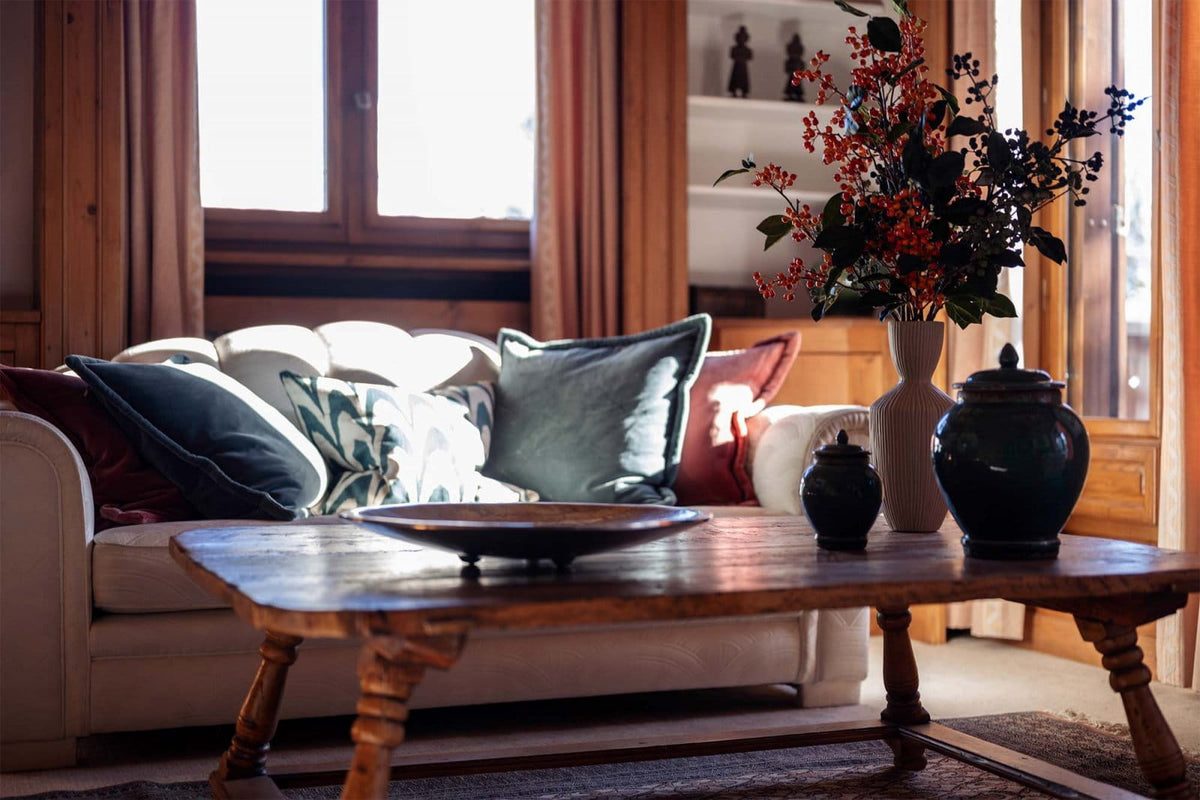 A cozy living room scene with a cream sofa adorned with dark blue and patterned throw pillows, set behind a rustic wooden coffee table. On the table sits a large black ceramic jar and a tall white vase filled with red and dark foliage.