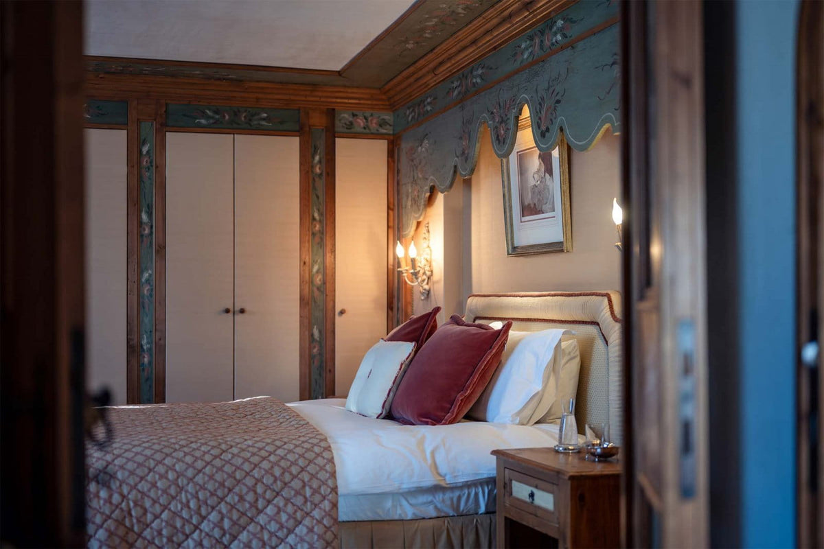 A cozy bedroom interior with a neatly made bed, white linens, burgundy and cream pillows, a patterned quilt, wooden nightstand, and ornate wall paneling with a blue-green valance.